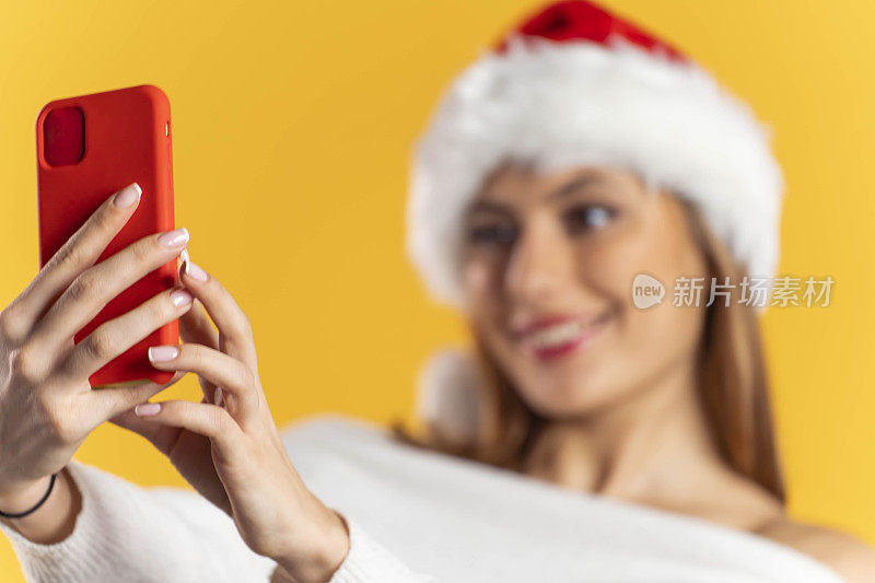 漂亮的年轻女子戴着圣诞老人帽，用手机/智能手机在黄色背景上视频通话/自拍